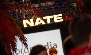 Beleuchtetes gelbes Schild mit dem Namen „NATE" über einer Bühne, davor Palmenblätter und Personen mit Headsets.