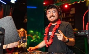 DJ mit Kopfhörern, Brille und rotem Blumenkranz an Mischpult, lächelt in die Kamera. Person im Hintergrund.