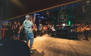 Person in Hawaiihemd steht auf Bühne vor Publikum in dunklem Club mit Holzboden und Neonbeleuchtung.