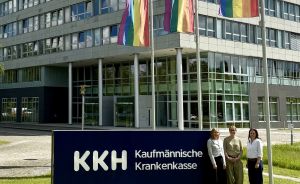 Drei Frauen vor dem KKH Bürogebäude, Regenbogenflaggen wehen im Hintergrund.
