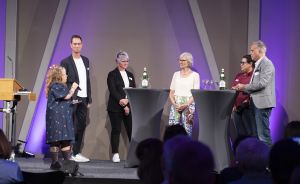 Sechs Personen auf der Bühne an Stehtischen mit Mikrofonen.