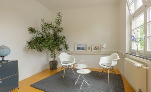 Helles Zimmer mit zwei weißen Schalenstühlen, rundem Beistelltisch, Grünpflanze, Bilderleiste und gelbem Holzboden.
