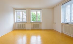 Leerer Raum mit gelbem Linoleumboden, weißen Wänden, zwei Fenstern mit Heizkörpern darunter und Jalousien.