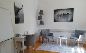 Helles Büro mit drei grauen Sesseln, weißem Tisch, Stehlampe, Whiteboard und zwei Wandbildern an weißen Wänden.