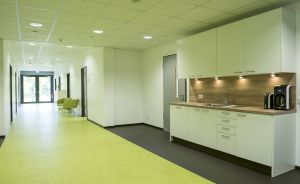 Heller Büroflur mit grünem Bodenbelag, weißer Küchenzeile mit Kaffeemaschine, Spüle und Oberschränken rechts.
