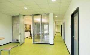 Büroflur mit grünem Boden, Glastrennwände mit Konferenzraum, weiße Decke mit Einbauleuchten, dunkle Türen rechts.