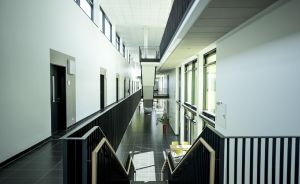 Moderner Flur mit schwarzen Geländern, weißen Wänden, dunklem Fliesenboden und Treppen, beleuchtet durch Fensterfronten.