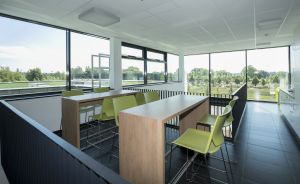 Heller Konferenzraum mit zwei Holztischen, grünen Stühlen, großen Fensterfronten und Blick auf Bäume und Wasser.
