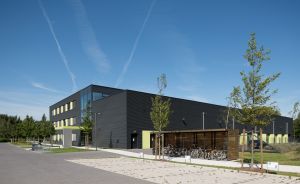 Modernes zweistöckiges Gebäude mit schwarzer Fassade und grünen Akzenten, Fahrradständer, junge Bäume, blauer Himmel.
