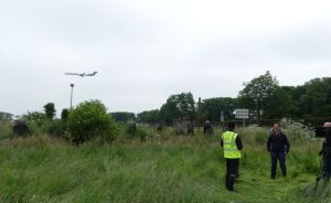 Drei Personen stehen auf einer Wiese, beobachten ein landendes Flugzeug. Eine trägt Warnweste, eine filmt mit Kamera.