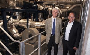 Zwei Männer in Businesskleidung stehen vor großen isolierten Rohrleitungen und technischen Anlagen in einem Industriegebäude.
