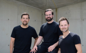 Drei Männer in schwarzen T-Shirts posieren lächelnd vor einer Betonwand.