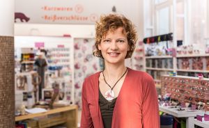 Frau mit lockigem Haar und roter Strickjacke steht lächelnd in einem Kurzwaren- und Reißverschluss-Geschäft.