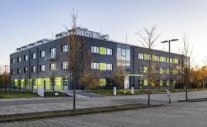Modernes 3-stöckiges Bürogebäude mit grünen Fensterelementen, Parkplatz, kahlen Bäumen, Abenddämmerung.