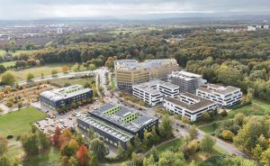 Luftaufnahme eines Gebäudekomplexes mit mehreren Bürogebäuden, umgeben von Bäumen und Grünflächen, Parkplätze sichtbar.