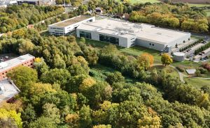 Luftaufnahme eines modernen Industriegebäudes mit Flachdach, umgeben von herbstlichem Mischwald und Parkplatz.