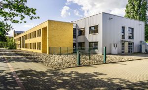 Zweistöckiges Gebäude mit gelber Klinkerfassade und grauer Metallverkleidung, Innenhof mit Pflastersteinen und Kiesbeeten.