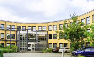 Gelbes, geschwungenes Bürogebäude mit Glaseingang, drei Etagen, Parkplatz mit Autos, umgeben von Bäumen.