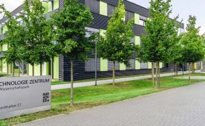Modernes Bürogebäude mit grün-schwarzer Fassade, Schild „Technologie Zentrum Hannover, Hollerithallee 17", Bäume, Rasen.