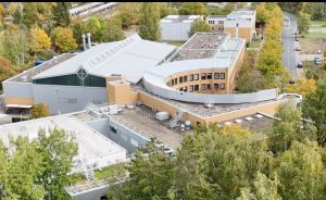 Luftaufnahme eines modernen Gebäudekomplexes mit geschwungener Fassade, umgeben von herbstlichen Laubbäumen und Parkplätzen.