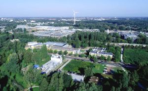 Luftaufnahme eines Industriegebiets mit Gebäuden, umgeben von Bäumen. Im Hintergrund ein Windrad und weitere Gebäude.