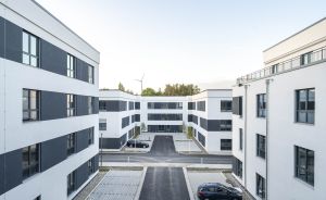 Moderner weißer Bürokomplex mit grau-weißer Fassade, Innenhof mit Parkplätzen und Autos, Windrad im Hintergrund.