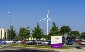 Firmengelände von Clarios mit Parkplatz, Windrad, Bäumen und weißem Schild auf blauem Himmel.