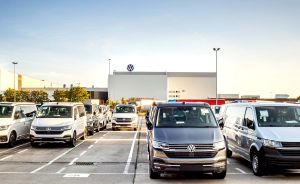 VW-Transporter und Vans parken in Reihen auf einem großen Parkplatz vor einem Volkswagen-Werksgebäude.