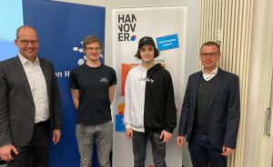 Vier Männer stehen vor einem Hannover-Banner. Zwei tragen Anzüge, zwei Jüngere sind casual gekleidet.