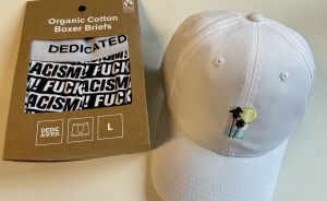 Schwarze Boxershorts mit „Fuck Racism"-Aufdruck in brauner Verpackung, Größe L, neben weißer Basecap mit Palmen-Stickerei.