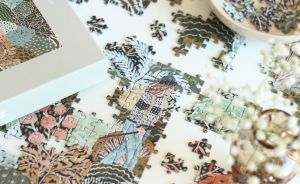 Puzzle wird auf weißem Tisch zusammengesetzt. Puzzlebox, Schüssel mit Teilen und Trockenblumen daneben.