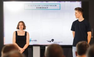 Zwei Personen stehen vor einer Leinwand mit der Aufschrift „Digitaler Livestream Demoday" vor Publikum.