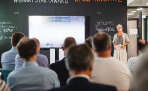 Frau spricht vor Publikum bei KPMG SmartStart Award Demoday. Leinwand und Kreidetafel mit Beschriftungen im Hintergrund.