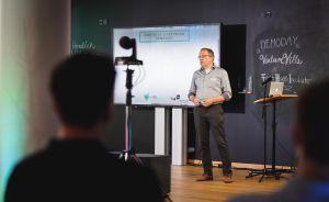 Mann präsentiert vor Leinwand mit "Digitaler Livestream Demoday" bei VentureVilla-Veranstaltung, Publikum im Vordergrund.