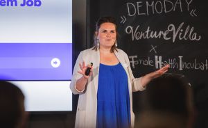 Frau in blauem Kleid präsentiert beim Demo Day von VentureVilla & Female Health Incubator vor Leinwand und Tafel.
