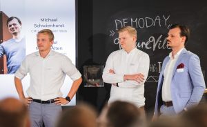 Drei Männer stehen vor einer Präsentationsleinwand und Kreidetafel mit Aufschrift „Demo Day" in einem Veranstaltungsraum.