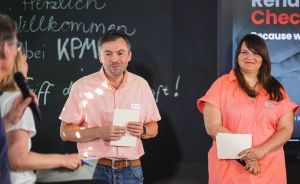 Zwei Personen in rosa Hemden halten Papiere bei einer Veranstaltung. Im Hintergrund Tafel mit "Herzlich Willkommen bei KPMG".