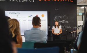 Frau präsentiert vor Publikum bei KPMG Innovate Demo Day. Hinter ihr Bildschirm mit "Mitgestalter"-Folie.