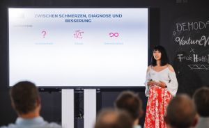 Frau präsentiert vor Publikum. Folie zeigt "Zwischen Schmerzen, Diagnose und Besserung" mit Symbolen für Unsicherheit, Chaos, Demotivation.