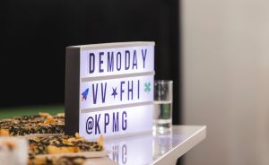 Beleuchtetes Leuchtkasten-Schild mit Text „DEMODAY VV FHI @KPMG" auf weißem Tisch, daneben Speisen und ein Glas.