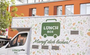 Weißer Food-Truck mit Logo „Lunch Box" und Slogan „Mampf dich lecker!", dekoriert mit Gemüse-Illustrationen.