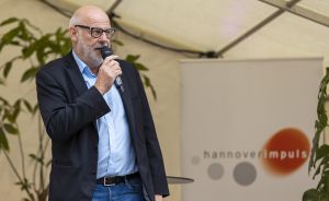 Mann mit Glatze und Brille spricht in Mikrofon vor hannoverimpuls-Banner unter Zeltdach.