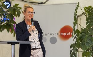 Frau mit Brille und Blazer spricht mit Mikrofon an einem Stehtisch vor einem „Hannover Impuls"-Banner.