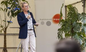 Frau mit Brille hält Mikrofon und spricht vor einem Hannover Impuls-Banner auf einer Veranstaltung.