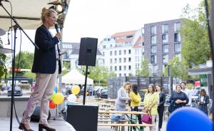 Frau spricht mit Mikrofon auf einer Bühne im Freien, Publikum steht dahinter, Luftballons und Gebäude im Hintergrund.