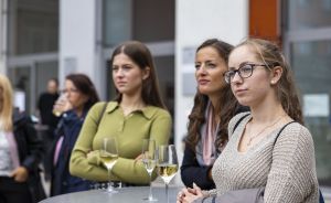 Drei Frauen stehen an einem Stehtisch mit zwei Weißweingläsern und schauen in dieselbe Richtung.