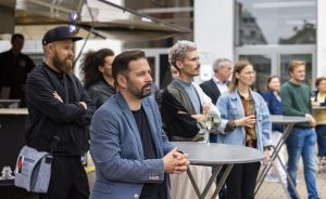 Gruppe von Menschen steht aufmerksam bei einer Outdoor-Veranstaltung, ein Mann im blauen Sakko lehnt an einem Stehtisch.