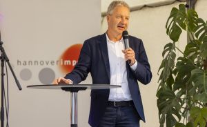 Mann im Blazer spricht mit Mikrofon an Stehtisch vor "hannoverimpuls"-Banner und Grünpflanze.