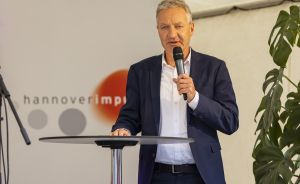 Mann im Blazer spricht mit Mikrofon an Stehtisch vor hannoverimpuls-Banner.
