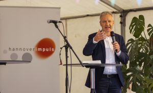 Mann im Blazer hält Mikrofon und spricht an Rednerpult vor hannoverimpuls-Banner unter Zeltdach.